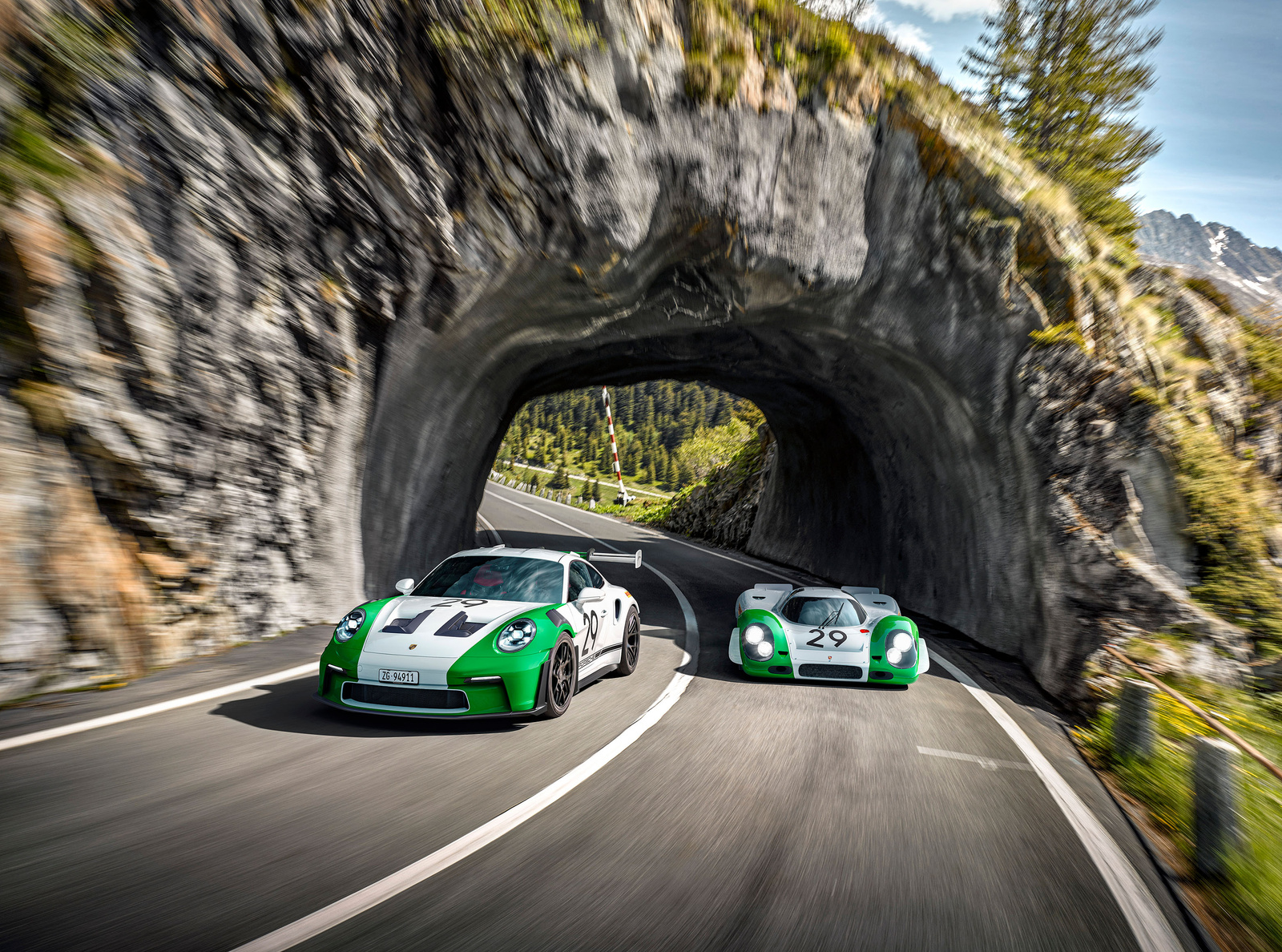 Zwei weiß-grüne Porsche 911 auf einer kurvigen Straße durch einen Felstunnel.