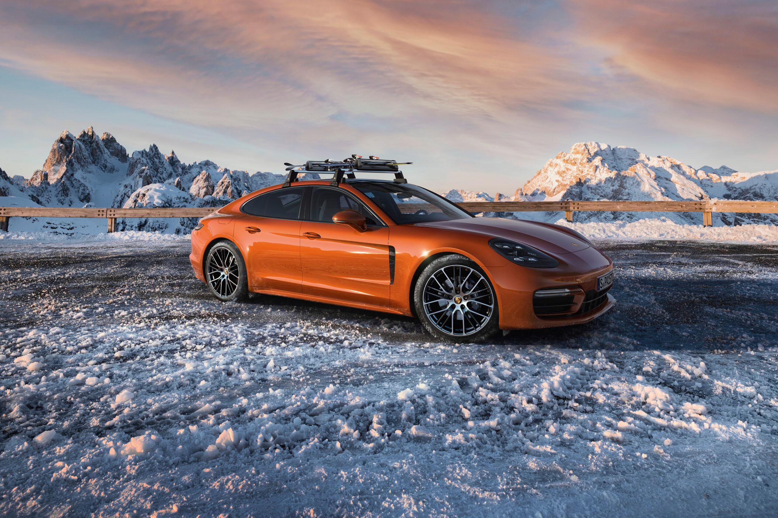 Orangefarbener Sportwagen mit Dachträger auf einer Schneefläche vor Bergen.
